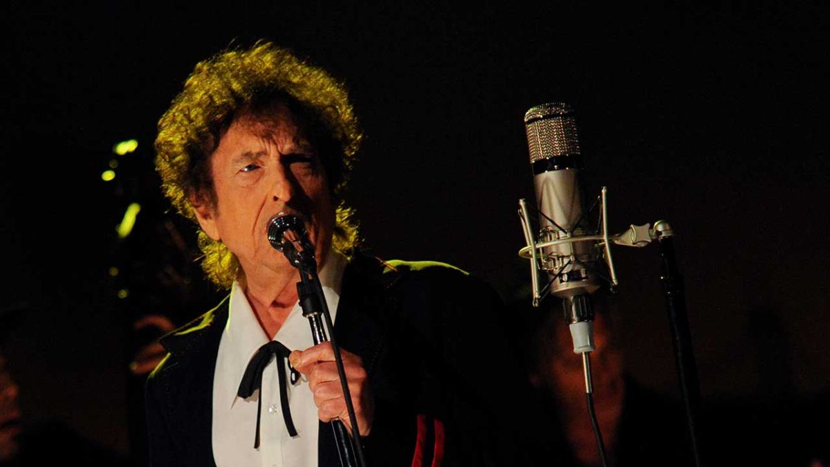 Bob Dylan mengenakan jas hitam, bernyanyi dengan mic di atas panggung